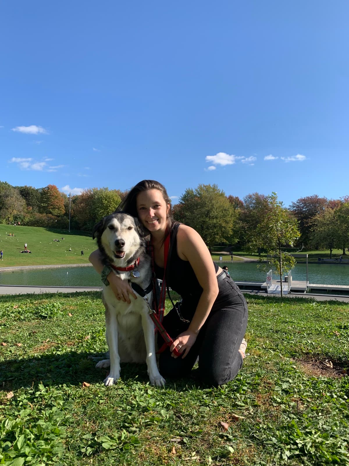 Liandra kneeling on the grass in a park, arm around her husky, with a lake and autumn trees behind them